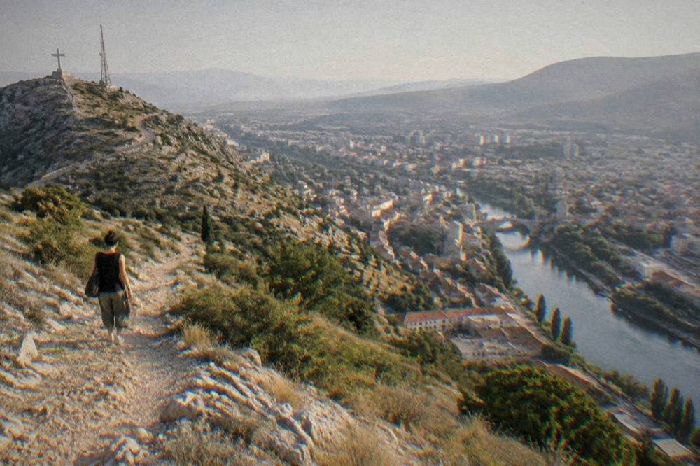 A person walking down a stony hill path toward a city with a river and many rooftops in late afternoon light