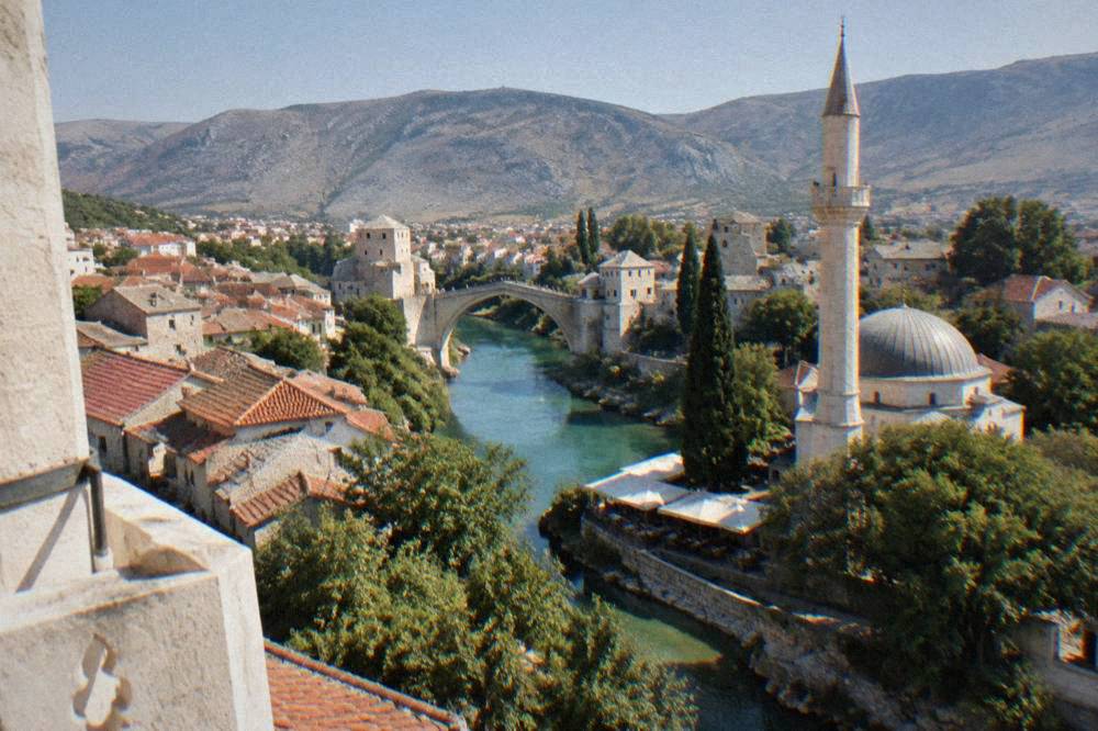 View from a minaret balcony over red rooftops toward a stone arch bridge spanning a turquoise river