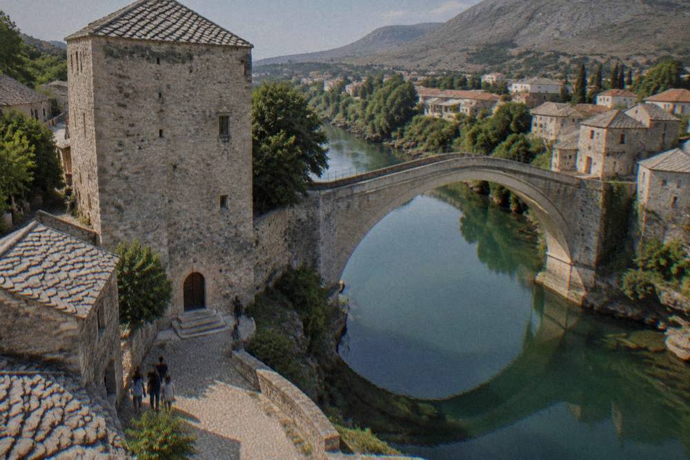 A stone bridge arch reflected in a green river between cobbled banks