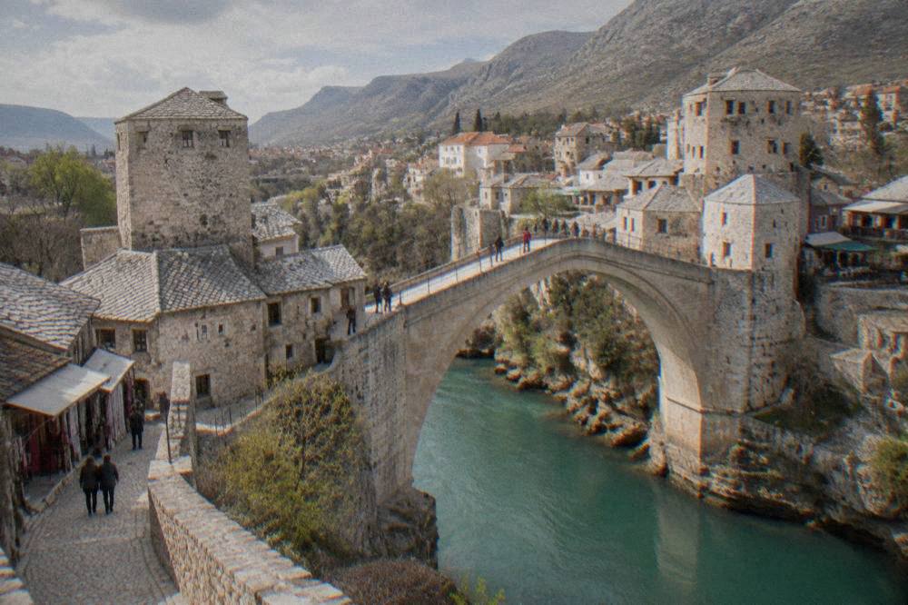 People crossing a pale stone bridge with cobbled streets and shop roofs behind them