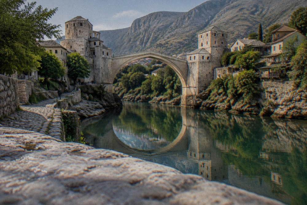 An old stone bridge reflected in still green water with hills behind