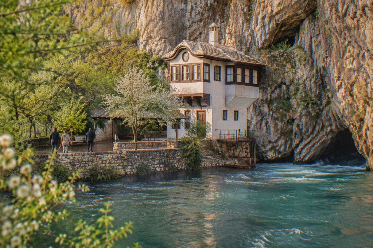 A white stone dervish lodge built into a cliff face above a turquoise river spring