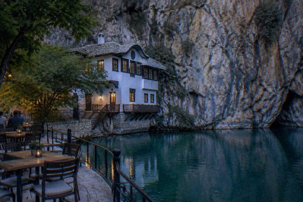 A riverside terrace with tables and chairs facing a tall limestone cliff