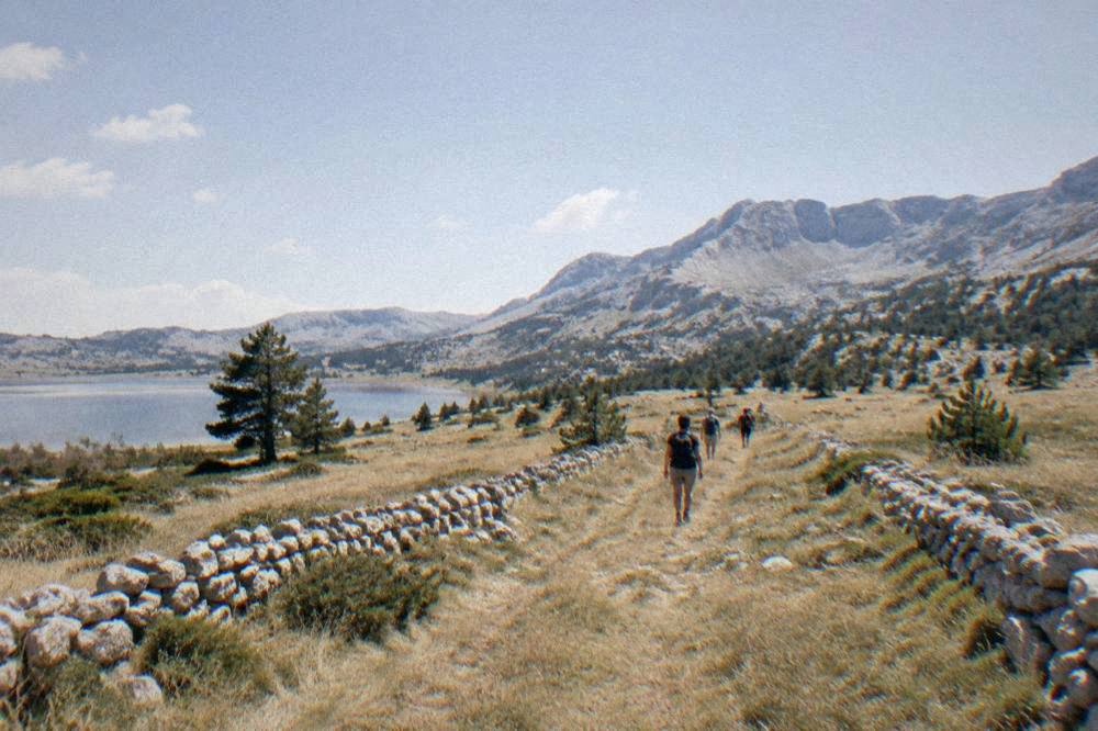 Hikers walking a grassy trail between dry stone walls toward rocky mountain peaks
