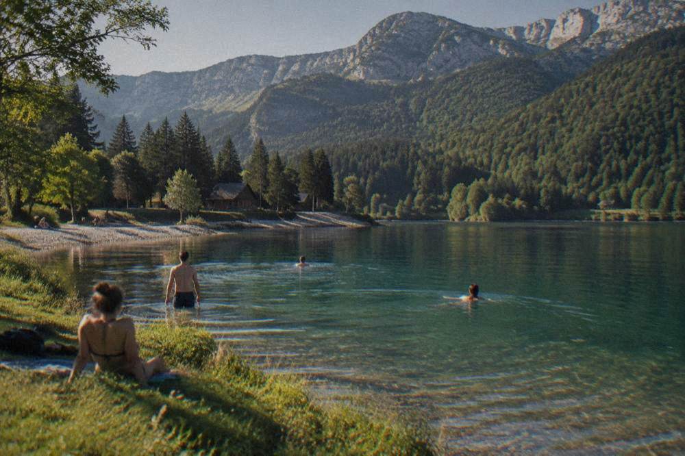 People swimming from a grassy bank into clear green lake water on a sunny day