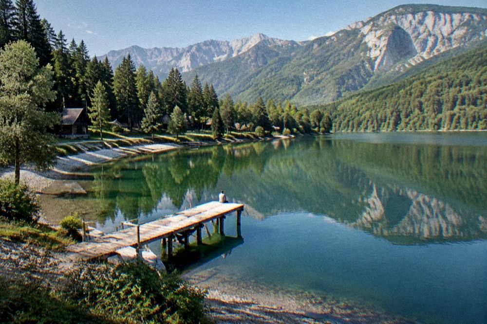 A wooden jetty over still green lake water with hills reflected in the surface