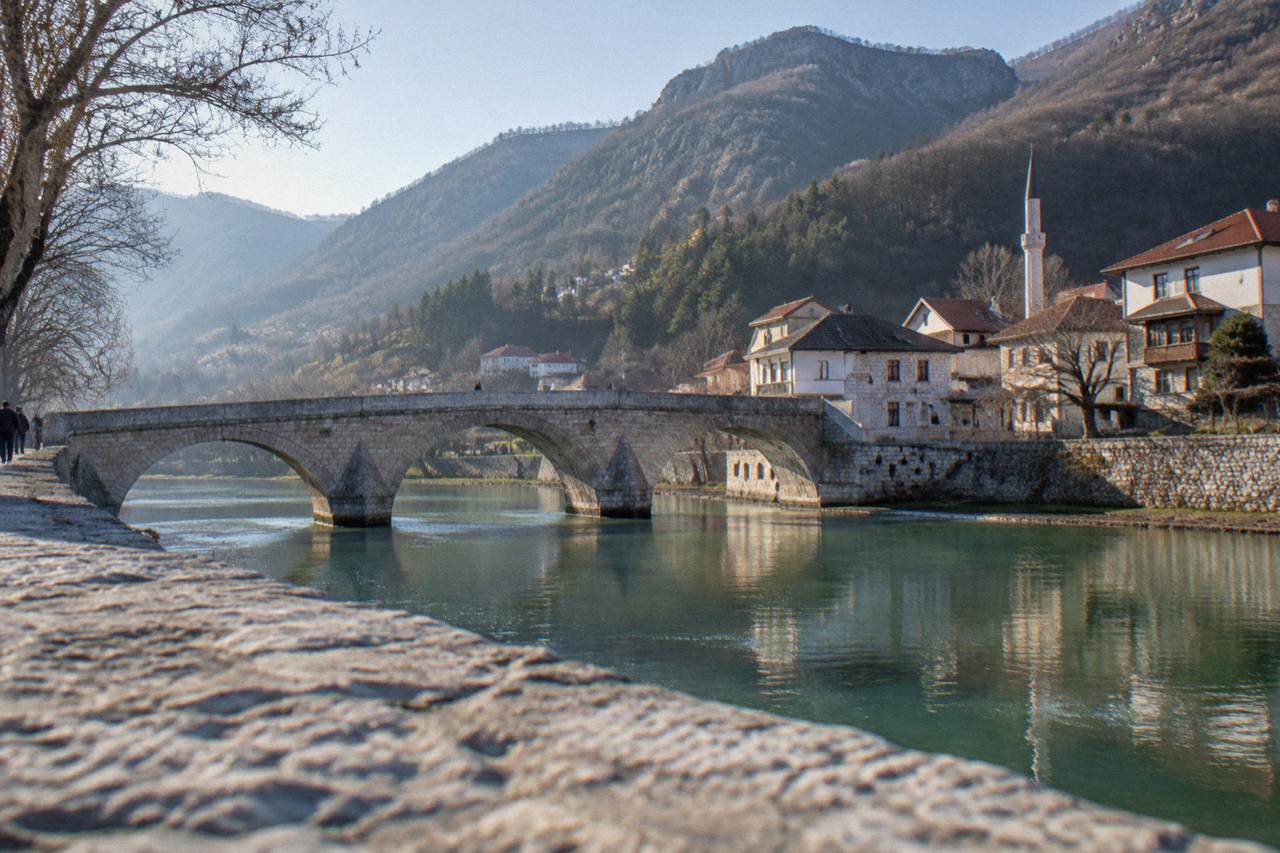 Stone bridge over a green river running through a small Bosnian town backed by wooded hills