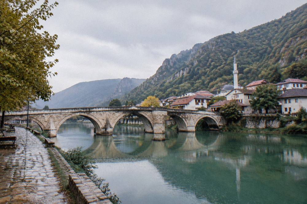 Multi-arched old stone bridge reflected in a calm green river at the edge of a small town