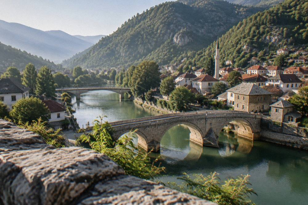Two stone bridges over a green river photographed from a hillside above a small town