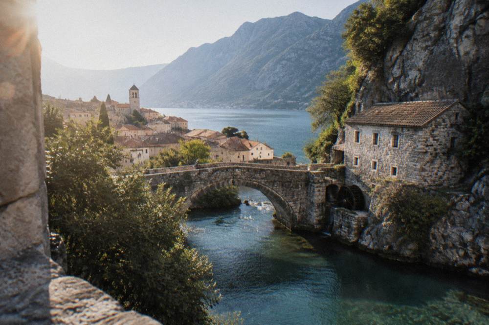 A stone bridge over a turquoise river beneath a cliff with a mill building at its base