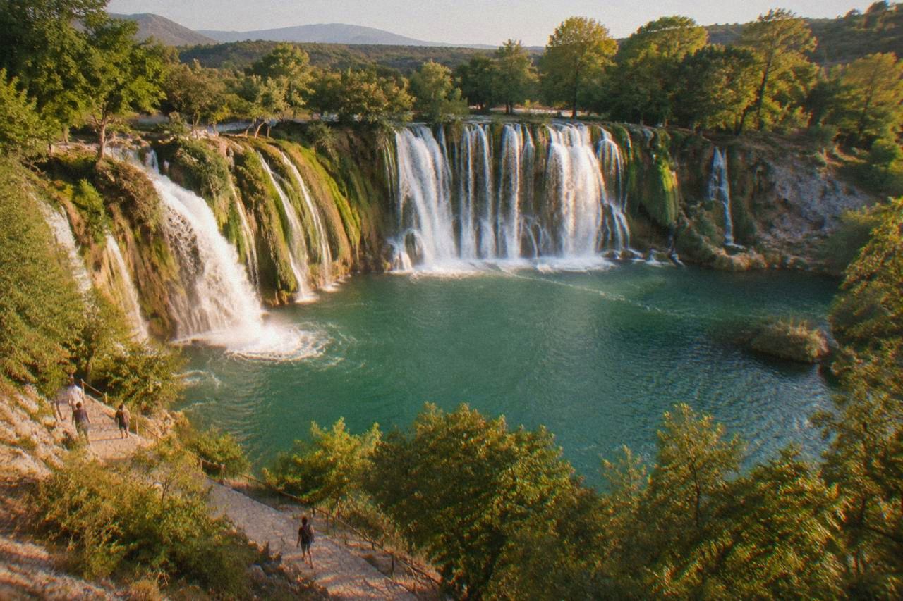 A wide waterfall curving across a rock face into a green pool surrounded by trees