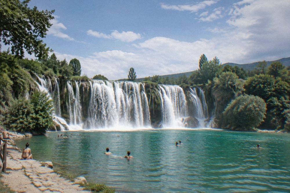 People swimming in the green pool below the wide waterfall on a sunny day