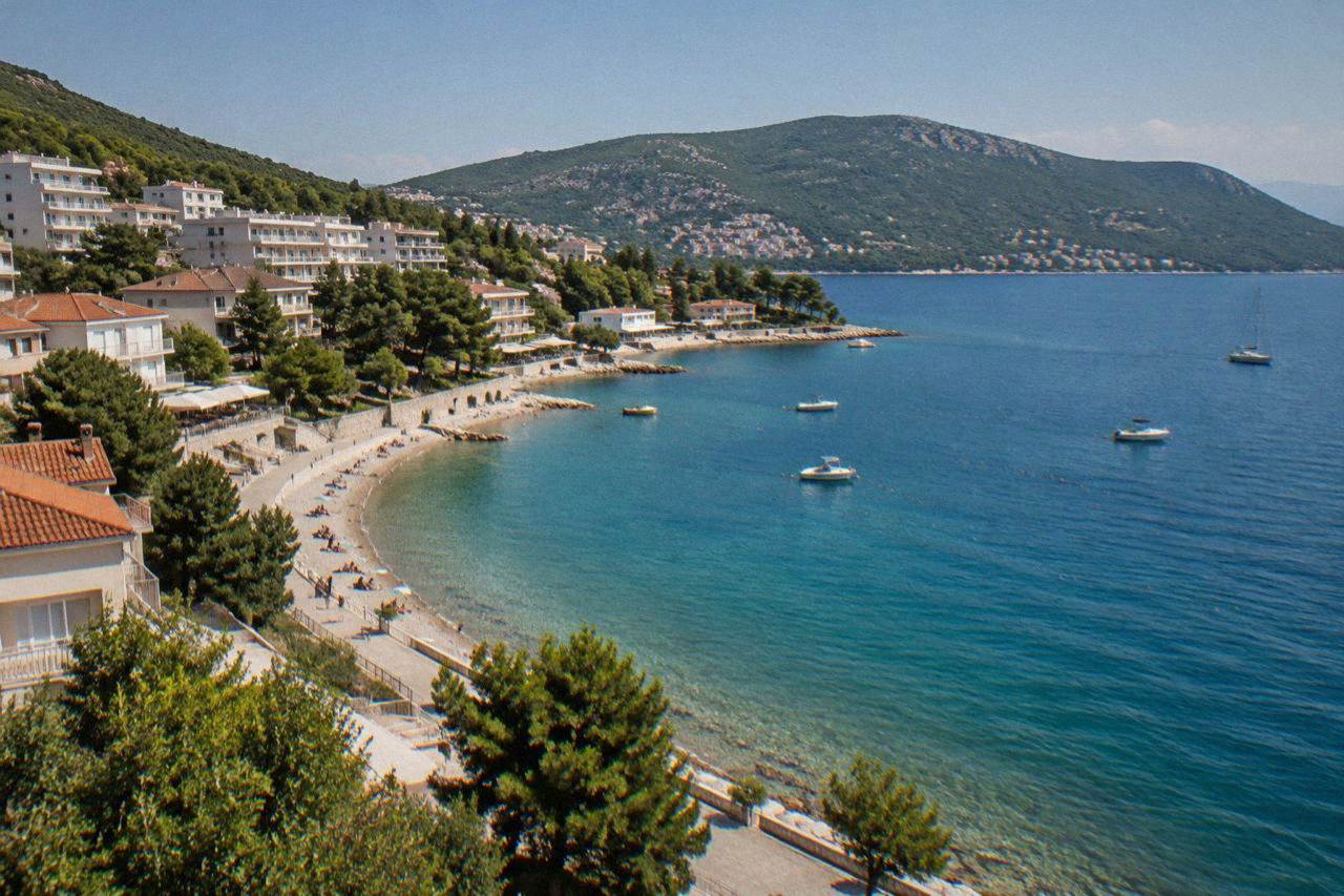 A pebble beach curving along a calm blue bay with low hills behind and small boats near the shore
