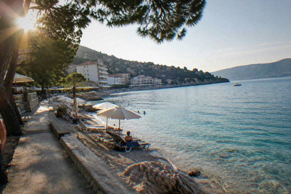 Sun umbrellas and loungers on a narrow pebble beach with clear turquoise shallows and pine trees