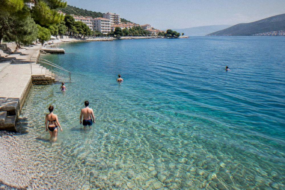 Calves and ankles of people wading into shallow clear water over pale pebbles on a sunny afternoon