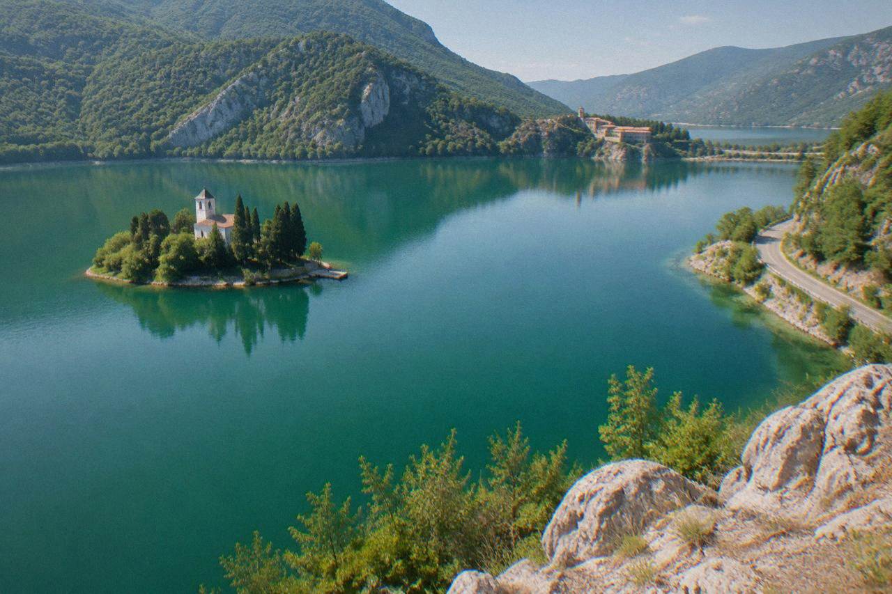 A calm green lake surrounded by wooded hills with a small peninsula monastery in the distance