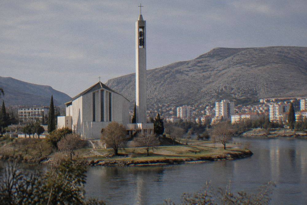 A white monastery church with a bell tower standing on a grassy peninsula between two arms of a lake