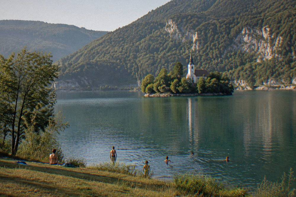 People swimming from a grassy lake shore with forested hills behind calm water