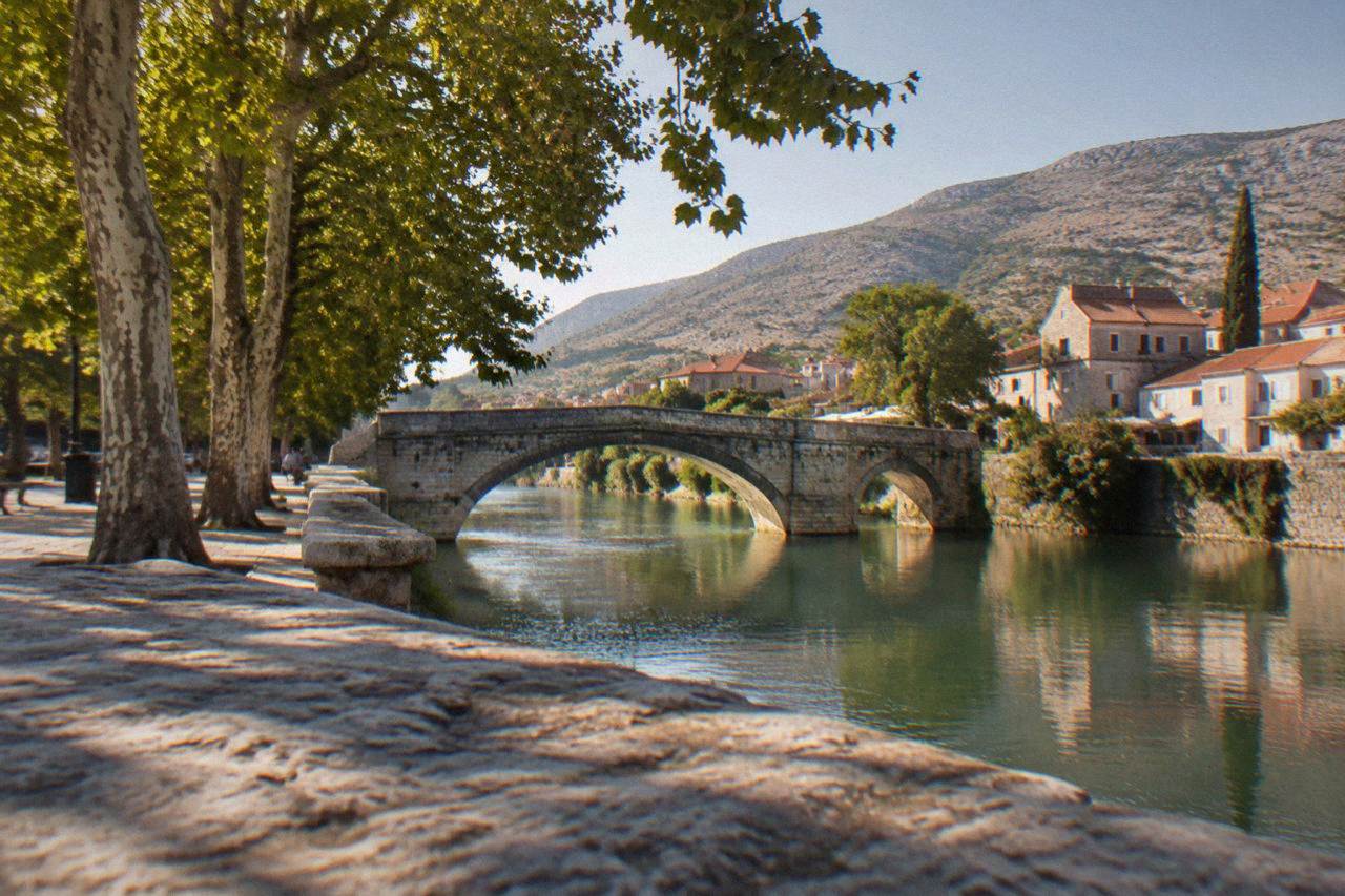 A stone bridge arching over a calm green river beside plane trees in Trebinje