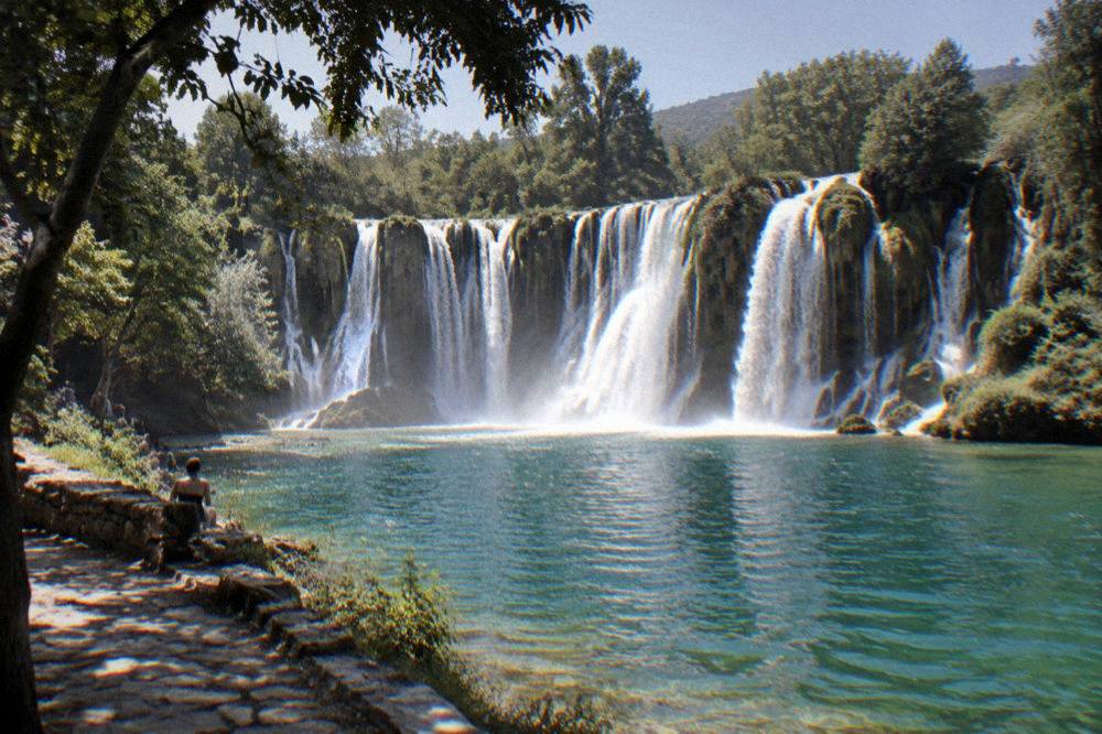 The wide cascade of Kravica waterfall falling into a green pool ringed by trees
