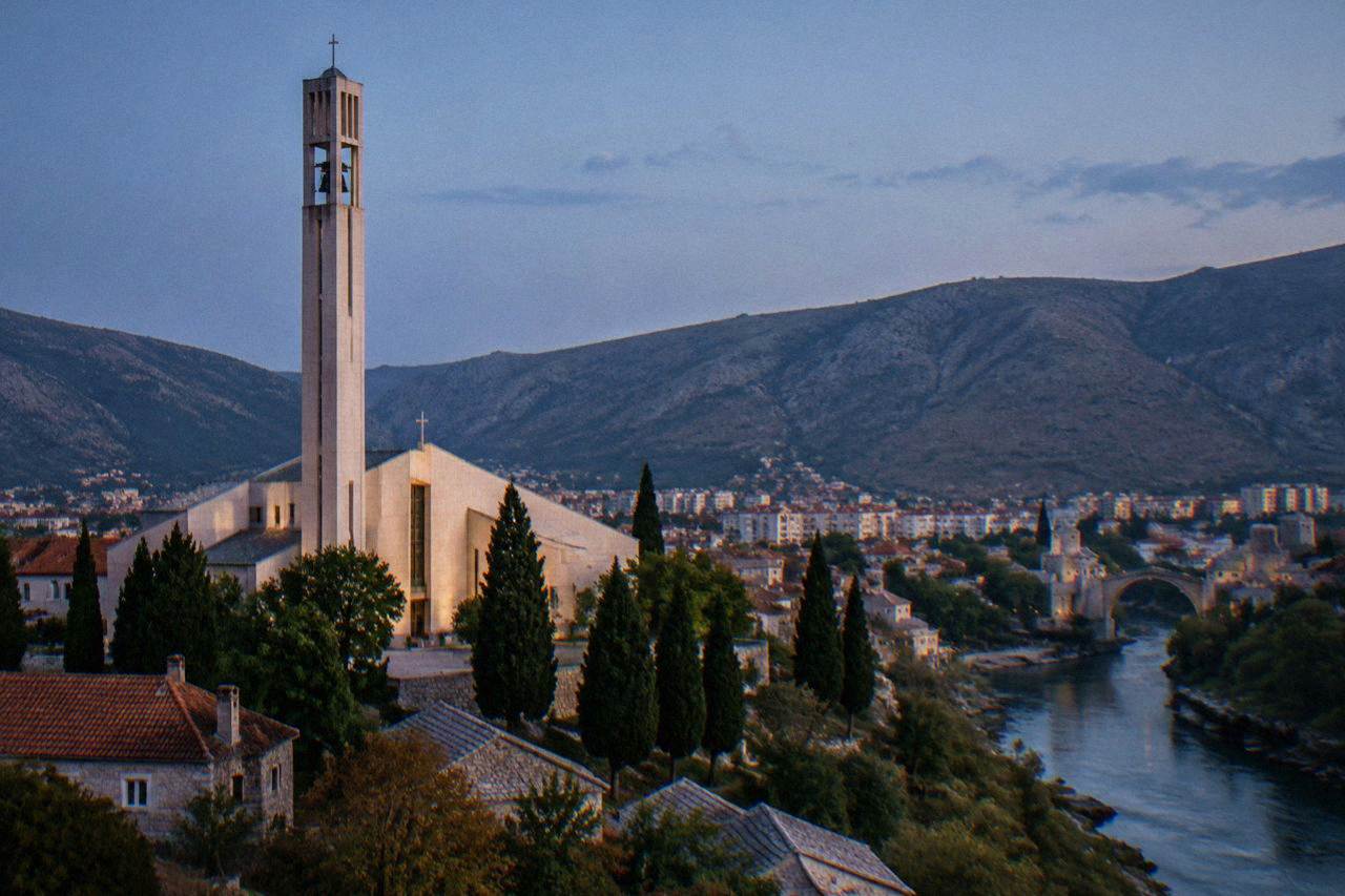 A stone Orthodox monastery church with a bell tower standing among cypress trees in the Herzegovina countryside
