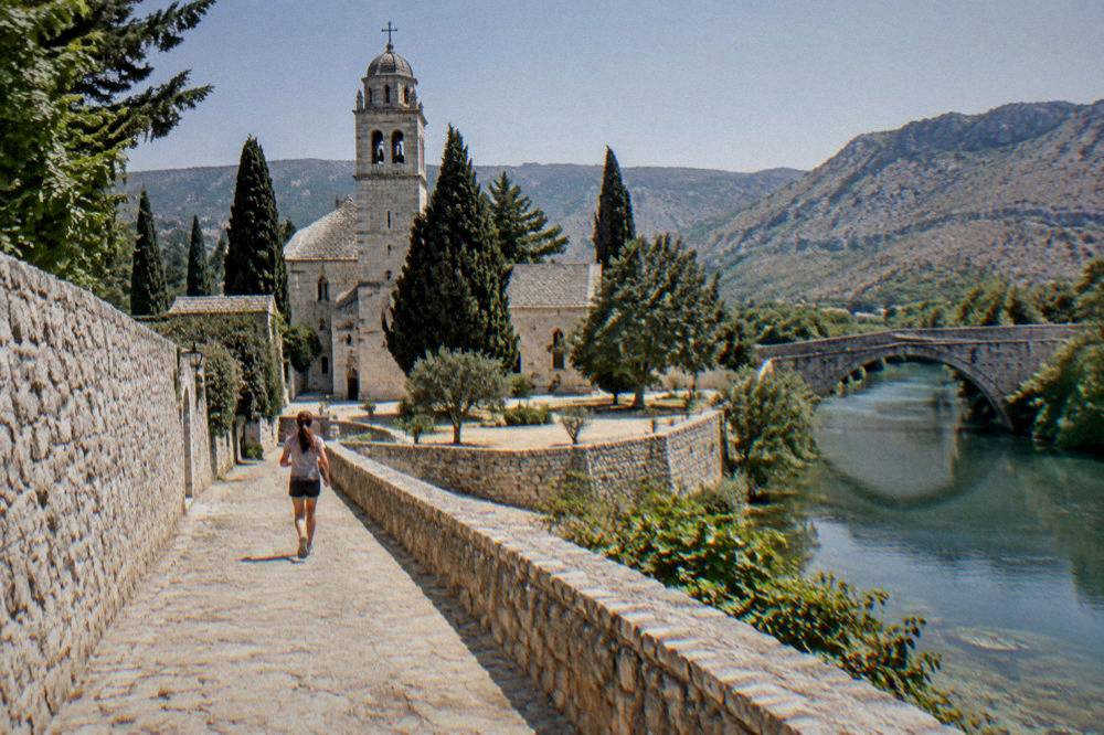 A stone bridge arching over the green Neretva river with hills rising behind it