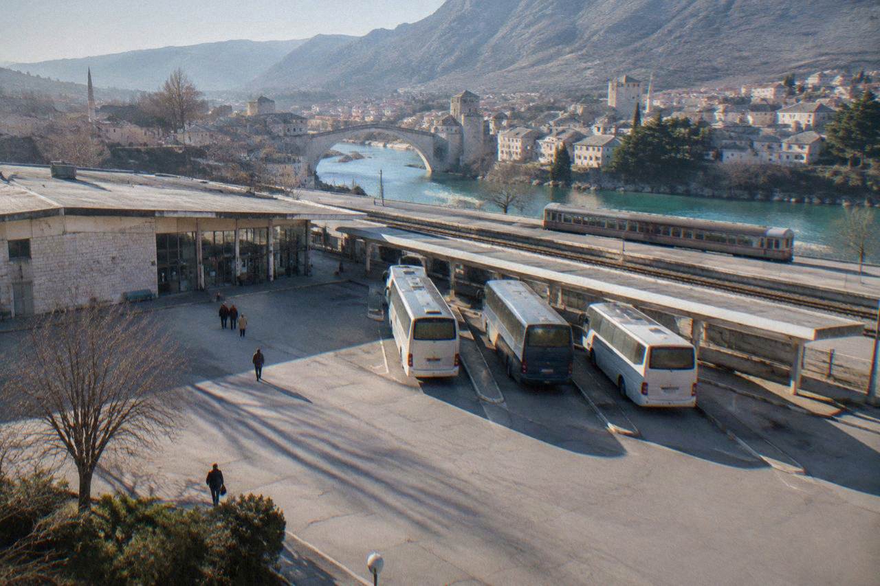 A stone bridge arching over a turquoise river in a Balkan city surrounded by hills