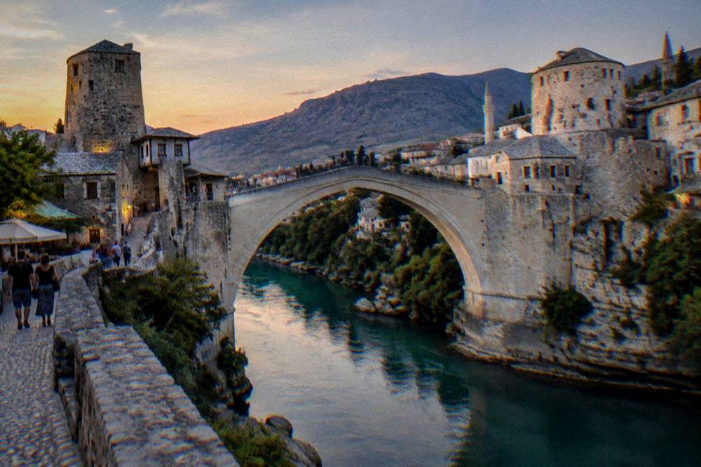 People strolling across the Old Bridge at sunset with hills behind the city
