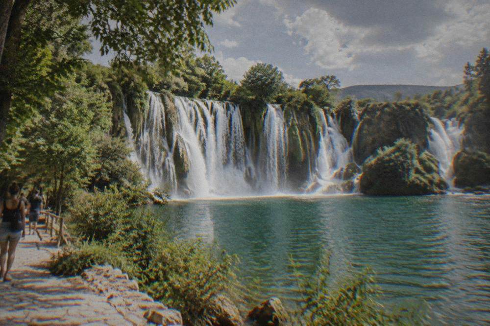 A wide waterfall curtain falling into a pool ringed by green trees at Kravica