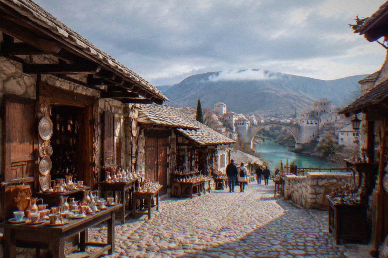 Families walking across a stone arch bridge above a turquoise river in a sunny old town