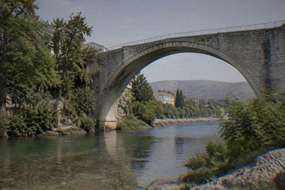 A stone bridge arch seen from a green river bank with shallow water below