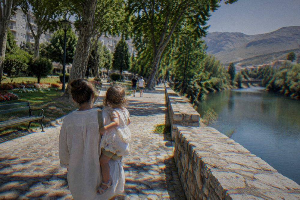 A parent carrying a toddler on stone cobbles beside a river embankment path